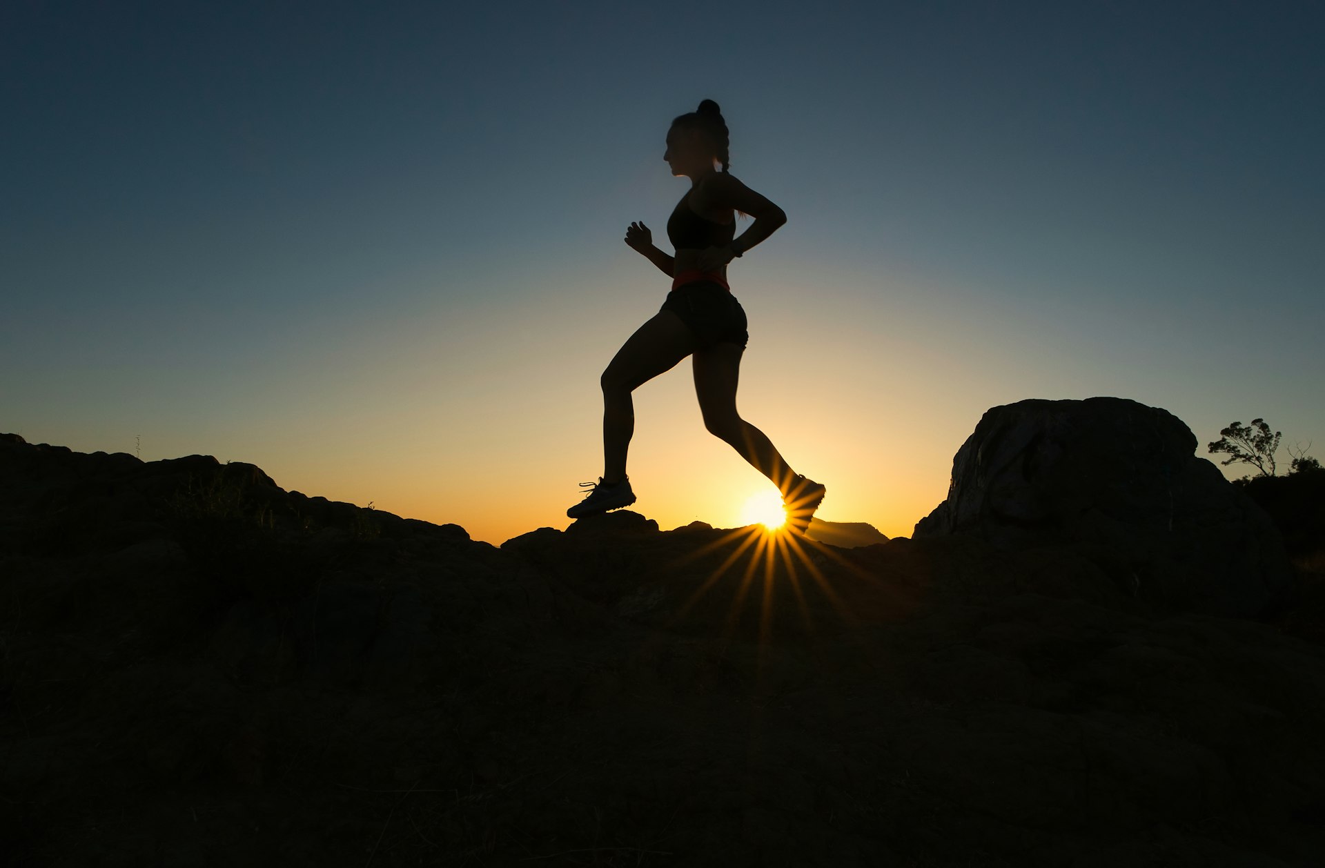 A woman running along a rocky ridge at sunrise, in silhouette against the sun