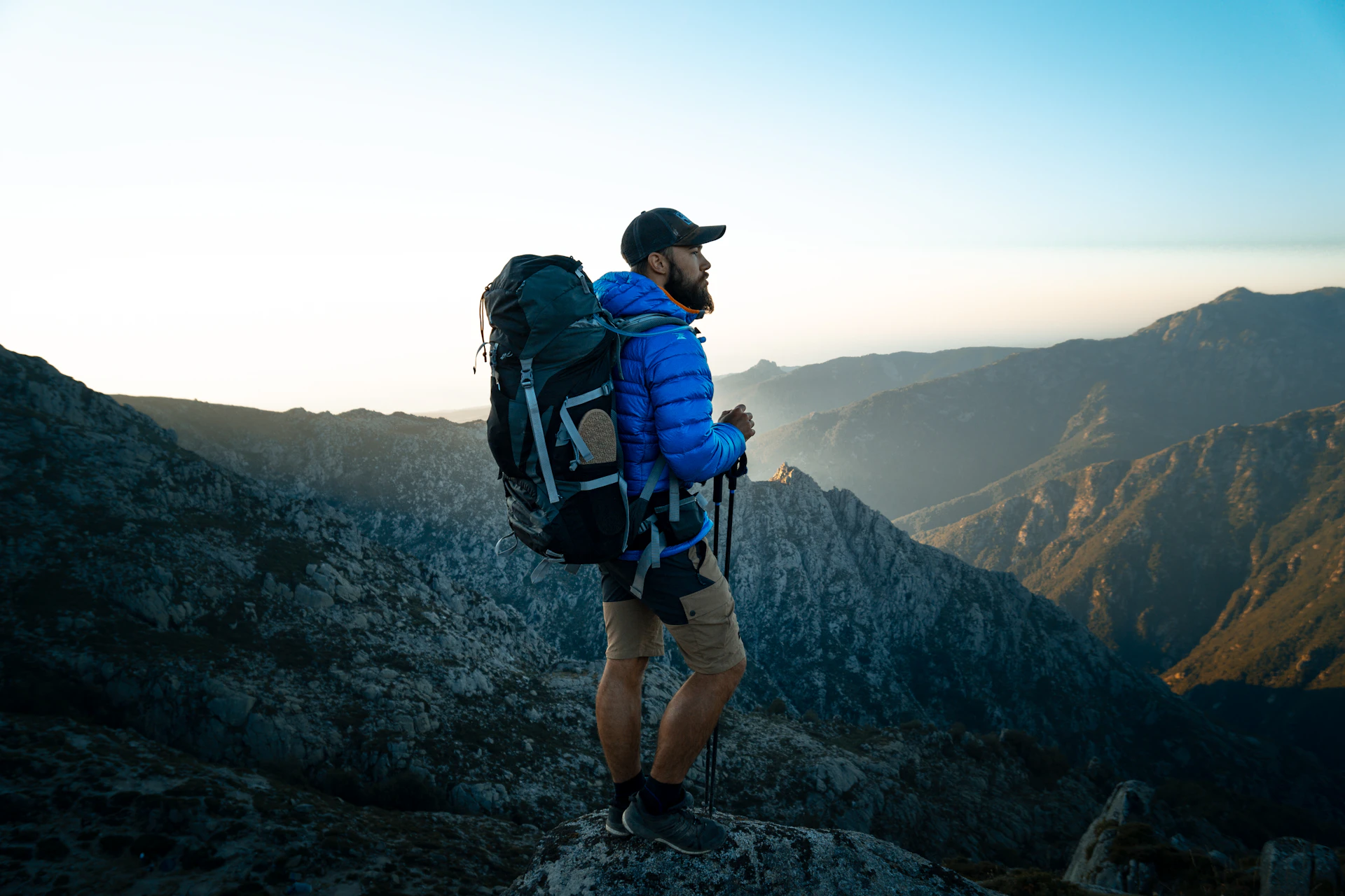 A trekker with a backpack and poles looking out over mountains at golden hour