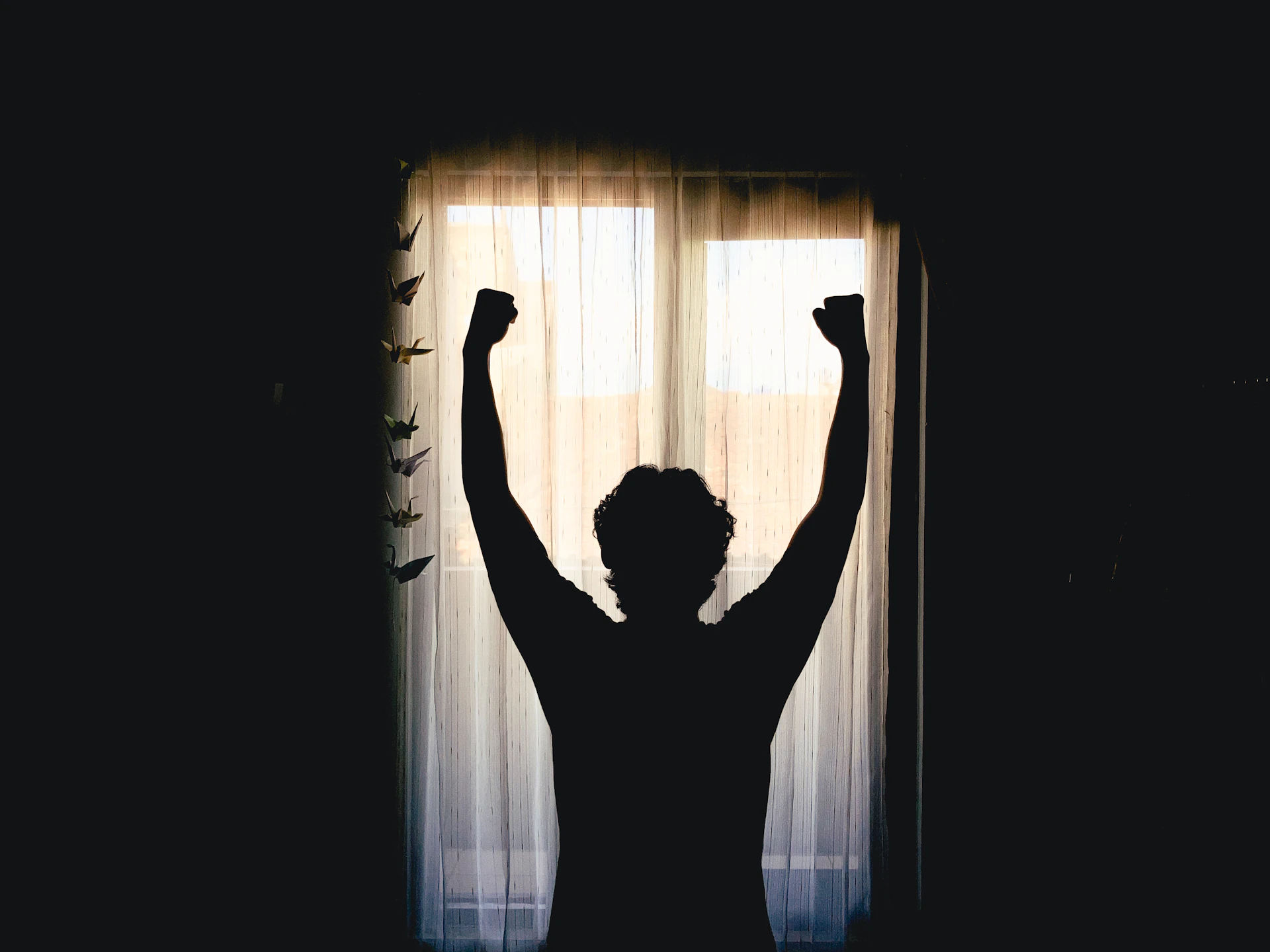 A person stretching their arms up in front of a bright window in the morning